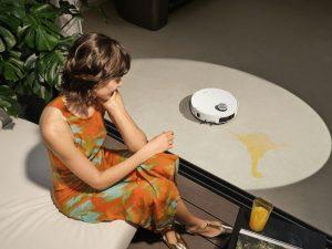 A woman in an orange dress relaxing on a sofa while a robot vacuum cleans a spill, illustrating Sola's concept of reclaiming free time.