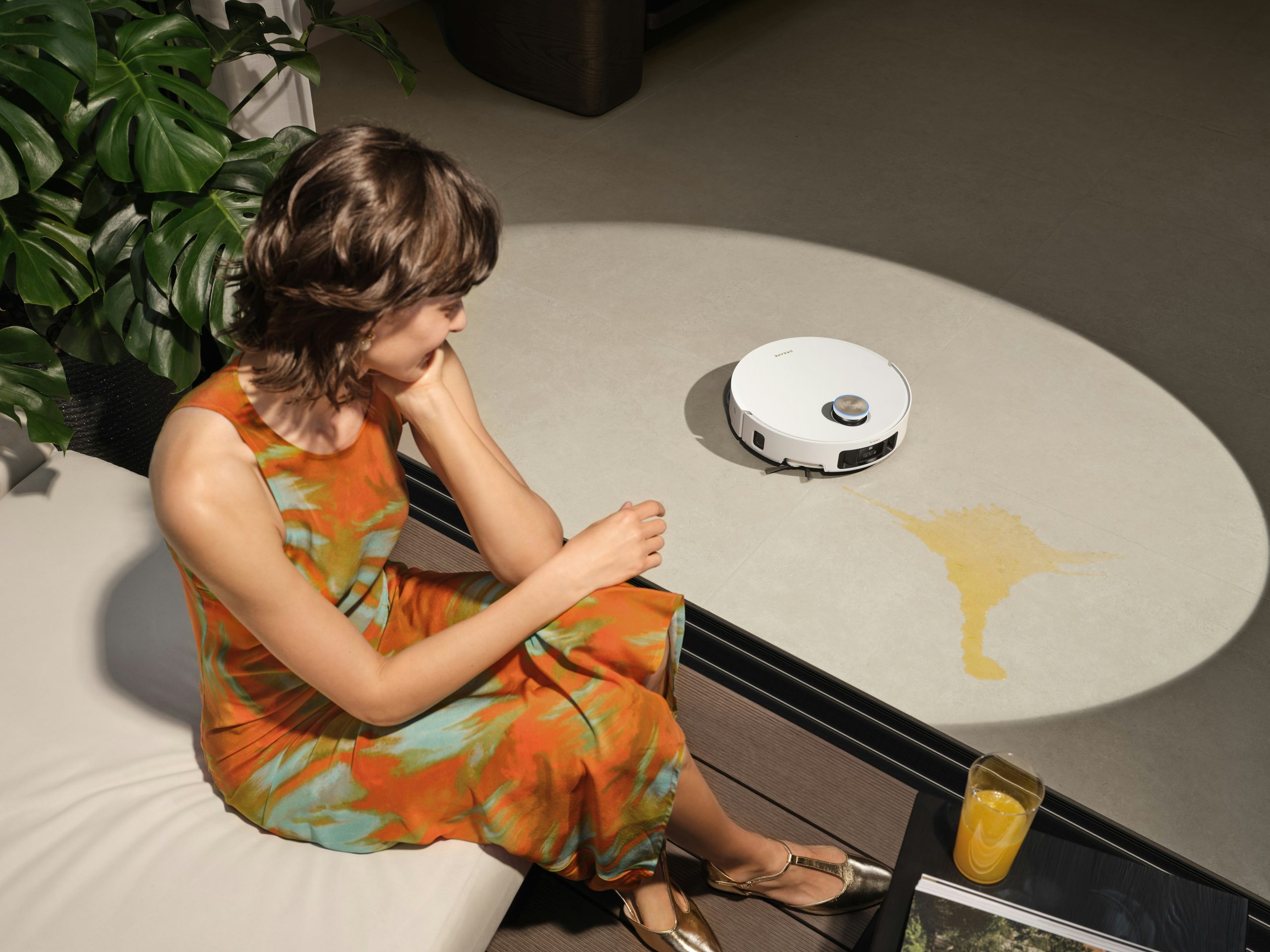 A woman in an orange dress relaxing on a sofa while a robot vacuum cleans a spill, illustrating Sola's concept of reclaiming free time.
